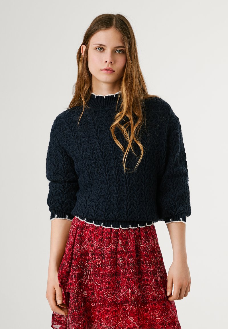 Jūras flotes krāsas adīts džemperis ar augstu apkakli un pūkainām piedurknēm; tam ir balta viļņota apakša, kas kombinēta ar sarkanu paisley raksta svārku.