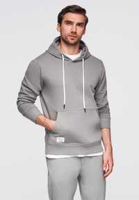 Šedá mikina s kapucí vyrobená z měkké tkaniny, s přední kapsou, bílými šňůrkami a žebrovanými manžetami. Nosená přes bílou košili.