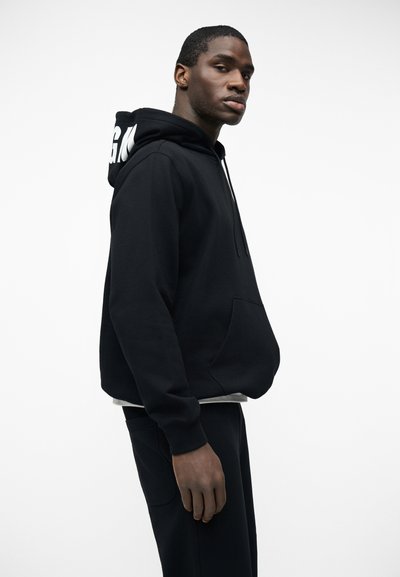 Jeune homme portant un sweat à capuche noir avec un texte blanc sur la capuche, debout de profil sur un fond blanc uni.