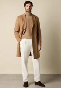 Mann steht vor einem einfarbigen beige Hintergrund und trägt einen camel Farbene Mantel über einem camel Farbene Rollkragenpullover, weiße pliierte Hose und dunkelbraune Mokassins, mit den Händen in den Taschen.