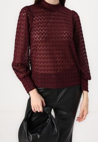 Bordeaux tekstureret bluse med høj hals og puffede ærmer, parret med sorte læderbukser og en sort taske.