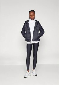 Naine seisab, seljas must puffer-jakk valge tõmblukuga pluusi, mustade retuuside ja valgete tossudega, lihtsa heleda tausta ees.