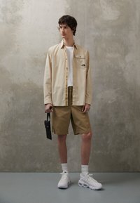 Camicetta beige con bottoni e tasca sul petto, pantaloni corti color sabbia e sneakers bianche. Il modello tiene una piccola borsa nera. Sfondo in cemento texturizzato.