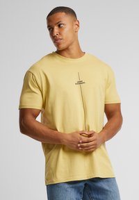 Camiseta de algodón amarilla de manga corta, de corte relajado, con un texto gráfico vertical en negro que dice "PIENSO DIFERENTE" en el frente.