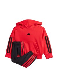 Czerwona bluza z kapturem z czarnym logo Adidas, z czarnymi paskami na rękawach. Czarne dresy z czerwonymi paskami i logo, wykonane z miękkiego materiału, w luźnym kroju.