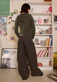 Zelený chlupatý svetr s kapucí, kombinovaný s širokými hnědými džíny. Pozadí zdobí police s různými knihami a dekorativními předměty.