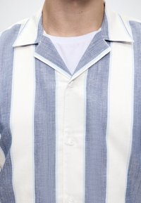 Mand iført en blå og hvid vertikalt stribet skjorte med knapper over en hvid t-shirt med rund hals.