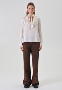 Blusa crema con scollatura arricciata e dettaglio con nodo, abbinata a pantaloni larghi marroni. La blusa ha maniche lunghe e una texture di tessuto liscia.