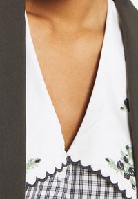 Vit skjorta med krage och broderade blommönster, lager-på-lager över ett mörkt rutigt tyg. Kragen har vågformade kanter. Mörk kavaj syns.