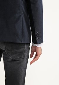 Uomo che indossa un blazer blu scuro con quattro bottoni sulla manica e jeans in denim neri, in piedi contro uno sfondo chiaro e uniforme.