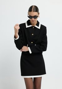 Schwarzes Blazerkleid mit weißem Kragen und Manschetten, ausgestattet mit goldenen Knöpfen. Das Material wirkt glatt, mit einer strukturierten Passform und minimalistischem Design.