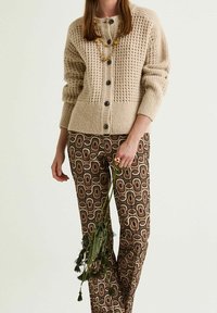 Cardigan beige lavorato a maglia con motivo a rilievo, con bottoni neri. Pantaloni marroni con motivo geometrico e un gambo verde.