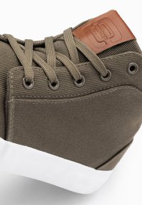 Zapatillas de lona verde oliva con parte superior texturizada, parche de cuero marrón y suela de goma blanca. Presentan ojales reforzados y cordones planos.
