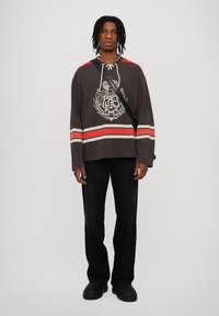 Camisa de hockey de manga larga en gris oscuro con rayas rojas y blancas, con un emblema gráfico en la parte frontal. Combinada con pantalones negros y zapatos negros.