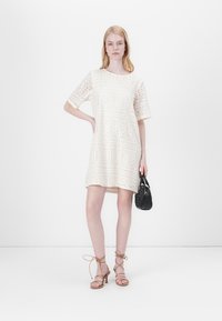 Abito in pizzo color off-white con maniche corte, motivi circolari e vestibilità ampia; abbinato a sandali beige con cinturini e una piccola borsa nera.