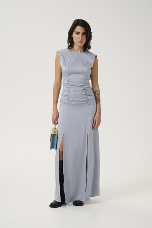 Light blue, sleeveless maxi dress with ruching, side slits, and smooth texture. Paired with a small handbag and black knee-high boots.