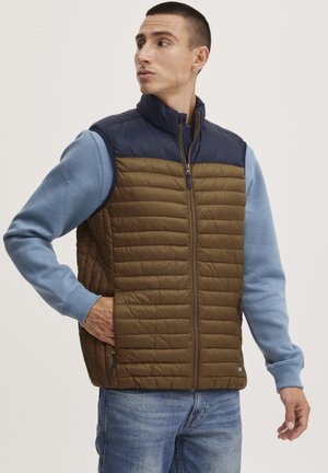 Wattierte Weste mit einem braunen gesteppten Design und einem hochgeschlossenen Marineblauen Kragen. Verfügt über Fronttaschen und einen Reißverschluss. Getragen über einem hellblauen Sweatshirt.