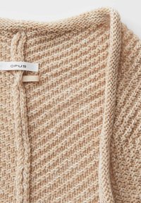 Gilet en tricot de couleur beige clair, présentant un motif texturé avec une légère brillance. Inclus une étiquette avec "OPUS" en noir.