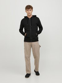 Zwarte rits-hoodie met een trekkoordcapuchon, gecombineerd met beige cargobroek en zwarte schoenen. Casual pasvorm met voorzakken en minimale details.