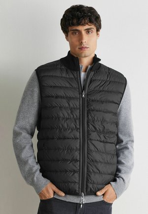 Młody mężczyzna w czarnej pikowanej kamizelce na zamek, noszący pod nią jasnoszary sweter z długim rękawem, stoi z rękami w kieszeniach na jednolitym tle.