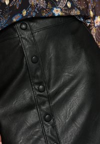 Gros plan d'une jupe en cuir noire avec une rangée de boutons recouverts et un haut à motif floral rentré au-dessus de la taille.