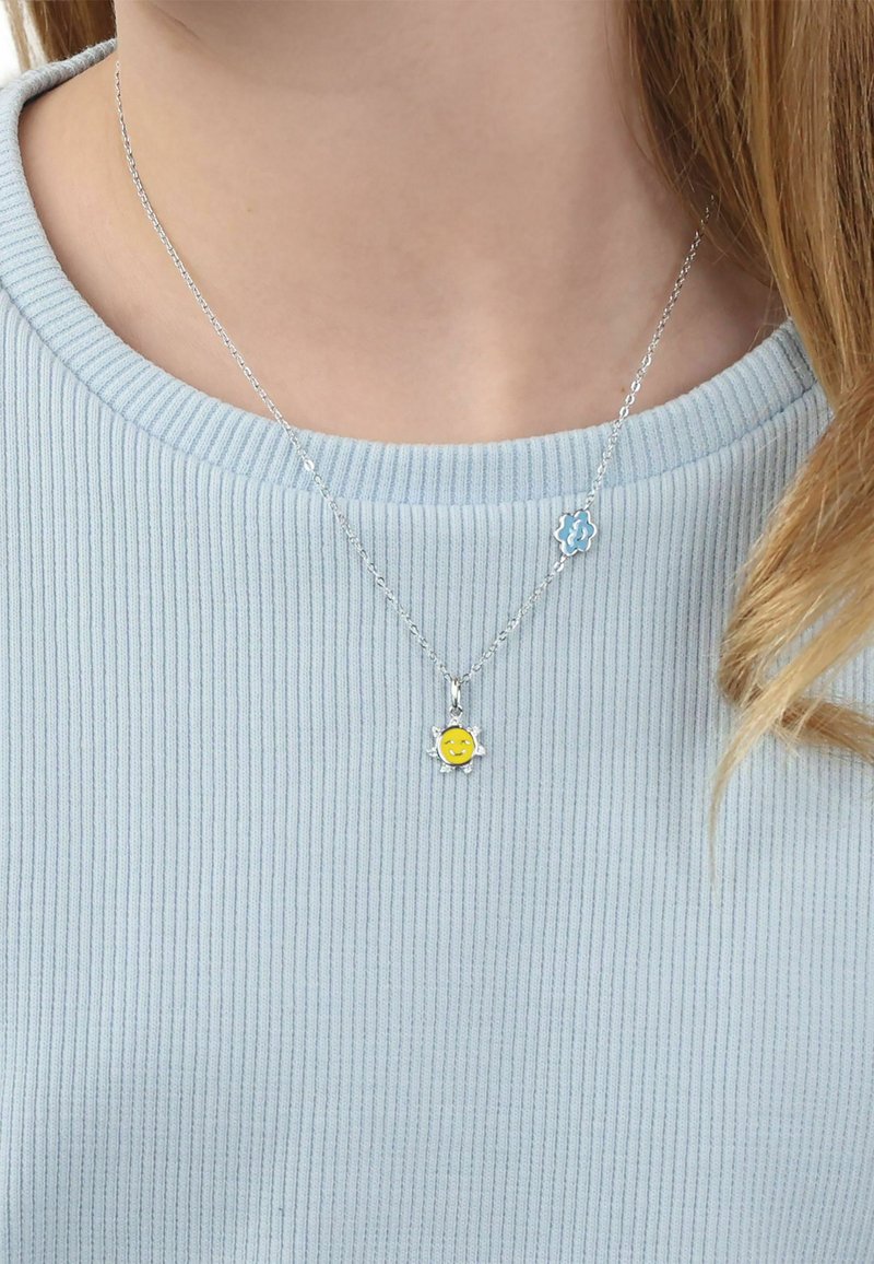Collana con catena d'argento con due ciondoli a forma di fiore, uno blu con una spirale bianca e uno giallo con un volto sorridente, indossata su una maglietta azzurro chiaro.
