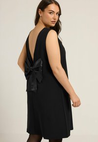 Vestido negro sin mangas con una espalda en V profunda y un gran lazo de satén en la cintura. Tejido liso con un corte holgado y un diseño minimalista.