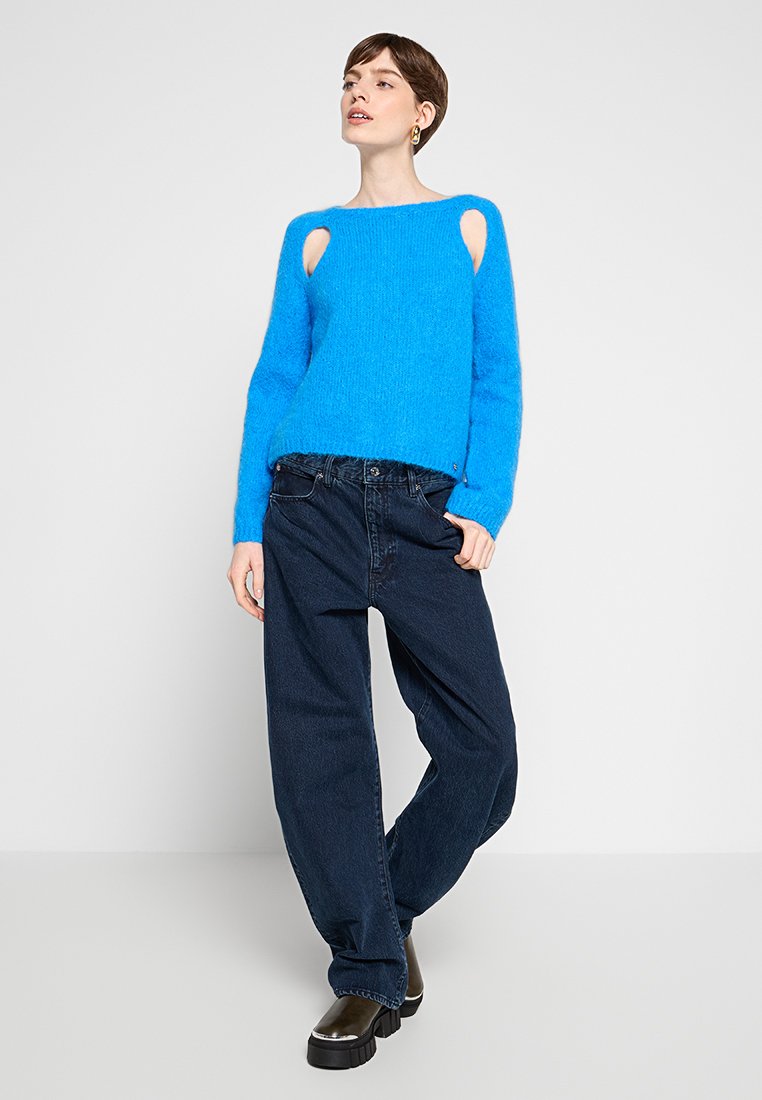 Maglione blu brillante e peloso con dettagli a taglio sulle spalle, abbinato a jeans di denim scuri e larghi e stivali neri robusti.