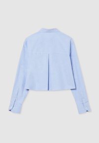 Chemise courte à manches longues bleu clair avec un pli dans le dos et des poignets boutonnés, présentée sur un fond neutre.