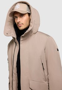 Chaqueta de plumas beige con una gran capucha, cierre frontal con cremallera y bolsillos laterales. Textura suave y diseño minimalista con acentos en negro.