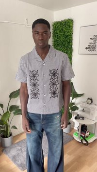 Camisa de manga curta em xadrez preto e branco com padrões florais bordados, usada com jeans de perna larga desbotados em azul, de pé no interior.