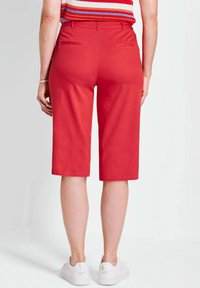 Shorts bermuda rouges en tissu lisse, dotés de deux poches arrière, d'une coupe droite et d'un design jusqu'aux genoux. Des baskets blanches sont portées.