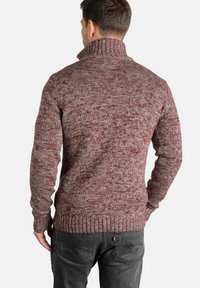Suéter de punto burdeos con cuello alto, patrón texturizado y puños acanalados. Lleva puesto sobre unos jeans oscuros. La tela parece cálida y gruesa.