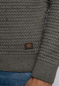 Suéter gris de punto con un diseño trenzado y texturizado, puños acanalados y un parche con el logotipo marrón en el dobladillo. El material parece grueso y acogedor.
