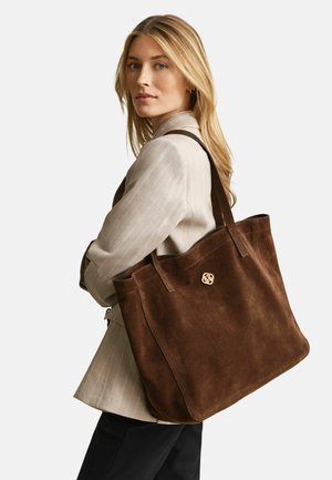 Blonde woman wearing beige blazer carries large brown suede tote bag with gold emblem over her shoulder, looking over her left shoulder.
