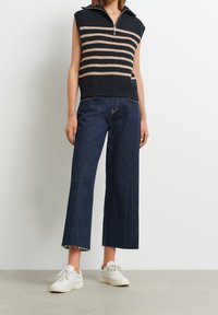 Marinblå och beige randig ärmlös stickad topp med dragkedja vid kragen, kombinerad med mörkblå vida jeans med fransade kanter och vita sneakers.