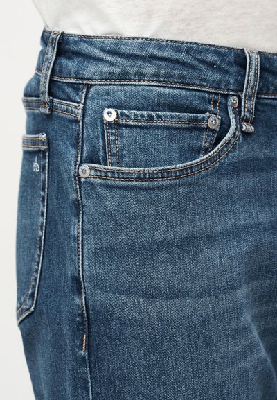 Gros plan sur la taille et la poche d'un jean en denim bleu avec des coutures visibles et des rivets argentés, porté avec un haut blanc.