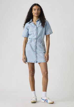 Frau mit langen lockigen Haaren, die ein hellblaues, jeansfarbenes Hemdkleid, weiße Socken und weiße Turnschuhe mit blauen und roten Streifen trägt.