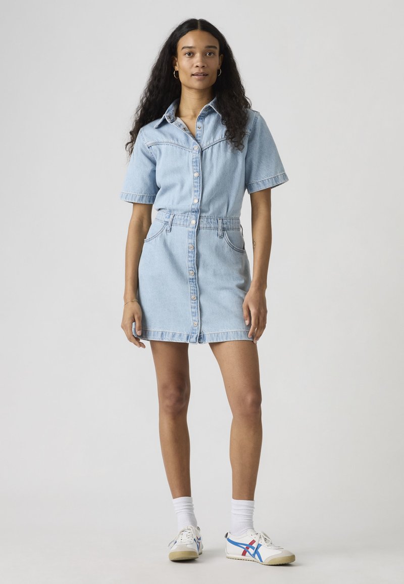 Femme aux longs cheveux bouclés portant une robe chemise en jean bleu clair, des chaussettes blanches et des baskets blanches avec des rayures bleues et rouges.