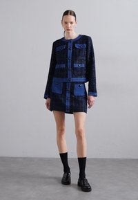 Set jakne in krila iz tweed materiala v temno modri barvi z kontrastnimi modrimi detajli. Ima gumbe, sprednje žepe in dolge rokave. Kombinirano z črnimi nogavicami in čevlji.