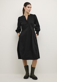 Črna A-linijska obleka z gumbasto zaklopko, dolgimi napihnjenimi rokavi, stranskimi žepi in gladko teksturo, v kombinaciji z debelimi zelenimi škornji.