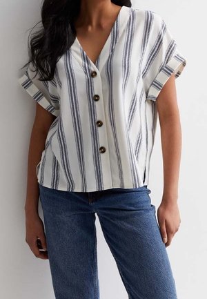 Kvinde iført en hvid kortærmet bluse med lodrette sorte striber og brune knapper, stylet med blå jeans.