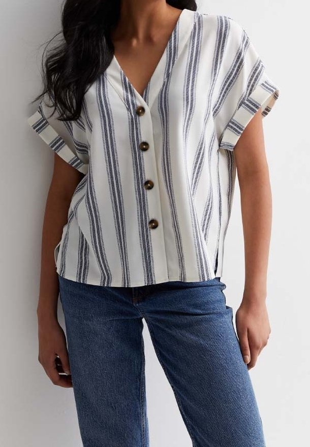 Femme portant un chemisier blanc à manches courtes avec des rayures noires verticales et des boutons marron, associé à un jean bleu.