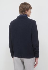Junger Mann, der seitlich steht, trägt einen marineblauen Strickpullover und hellgraue Hose vor einem schlichten, hellen Hintergrund.