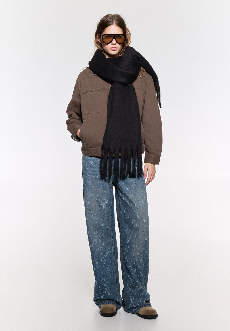 Giacca marrone oversize, sciarpa di lana nera con frange, jeans azzurri chiari con macchie di vernice e scarpe slip-on beige.