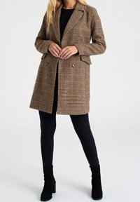 Brun rutigt kappa med notch-krage, dubbelknäppning och sidofickor. Bärs över en svart topp och matchas med svarta tajta jeans.