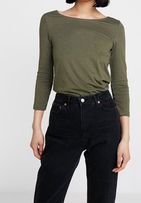 Maglietta aderente a maniche lunghe verde oliva con scollatura ampia, abbinata a jeans neri a vita alta con dettagli in rame e cinque tasche.