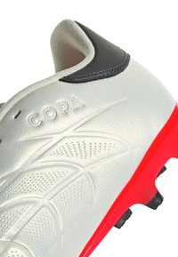 Botín de fútbol con un upper de cuero plateado, acento negro cerca del talón, superficie texturizada y una suela roja vibrante con tacos negros.