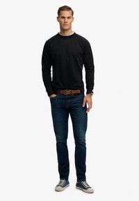 Felpa nera con collo rotondo, abbinata a jeans in denim blu. Cintura marrone con fibbia rettangolare e sneakers blu con dettagli bianchi.