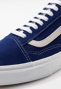 Baskets en daim bleu avec lacets blancs et une bande latérale beige, dotées de détails de couture et d'une semelle en caoutchouc texturée.